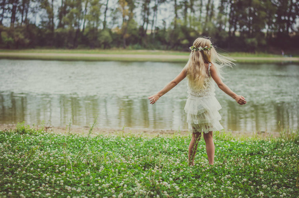 niña con el pelo largo y rubio en vestido blanco bailando por la noche en la hierba verde junto al río
 - Foto, imagen