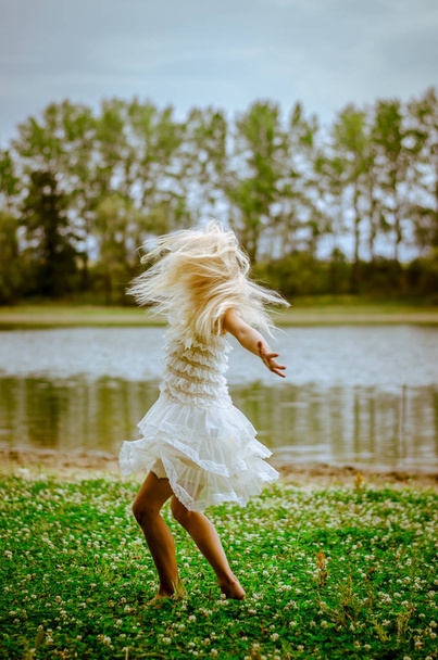 beautiful caucasian girl with long blond hair in colorful dress dancing wild in the green grass by the river - Фото, изображение