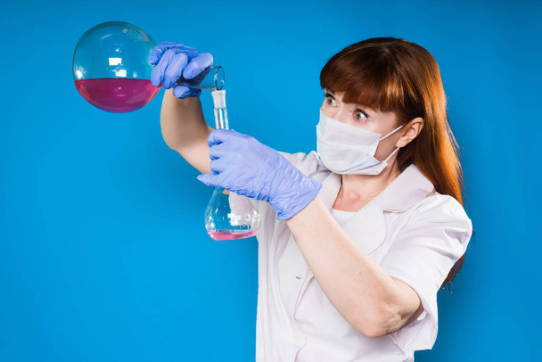surprised adult woman chemist in a white protective mask mixes chemical solutions in flasks - 写真・画像