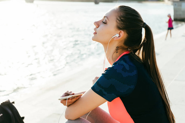 een aantrekkelijke jonge meisje in sportkleding is rust na scholing, luisteren naar muziek in hoofdtelefoons, kijken naar de rivier - Foto, afbeelding