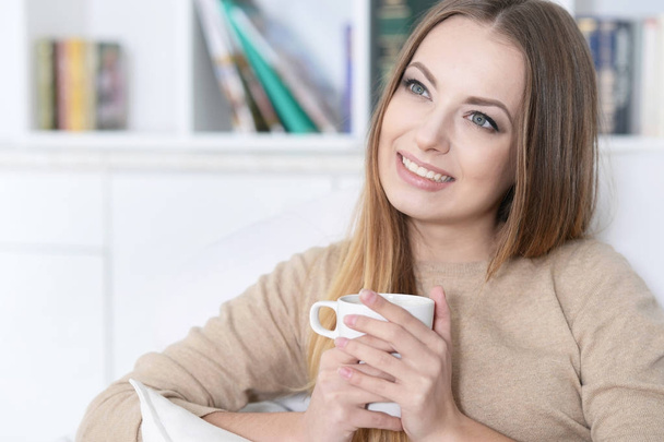  Młoda piękna kobieta z filiżanką herbaty na kanapie w domu - Zdjęcie, obraz