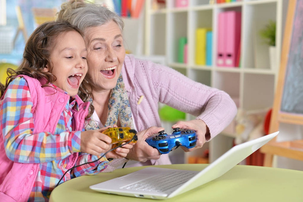 granny with her granddaughter playing computer game on laptop at home - Фото, изображение
