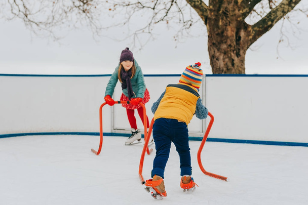 Щасливі смішні маленькі діти практикують з підтримкою катання на ковзанах, носять яскраві капелюхи та теплі куртки. Зимова діяльність для дітей
 - Фото, зображення