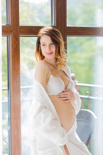 Fiatal terhes nő a fehér fehérnemű álló ablak közelében - Fotó, kép