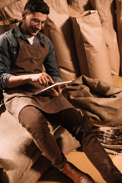 улыбающийся работник в фартуке с помощью планшета, сидя на мешок
 - Фото, изображение