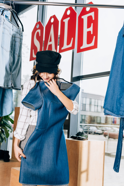happy young woman looking at blue denim dress at clothing store - Foto, imagen