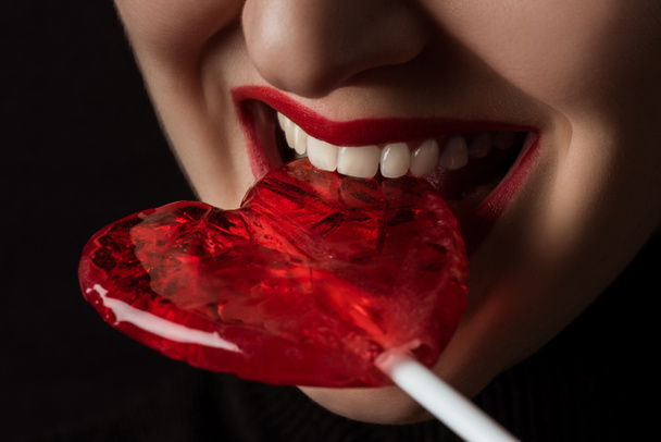 abgeschnittenes Bild einer Frau, die herzförmigen Lutscher beißt, isoliert auf schwarzem, Valentinstag-Konzept  - Foto, Bild