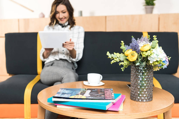 šťastná mladá žena pomocí tabletu, zatímco sedí na gauči v kavárně s květinami a časopisy na stole - Fotografie, Obrázek
