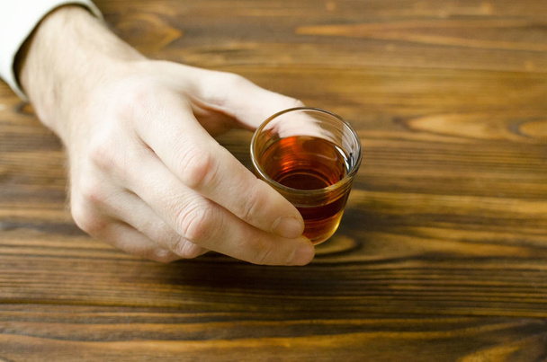 Держите в руке маленький стакан с алкогольным напитком на деревянном столе
 - Фото, изображение