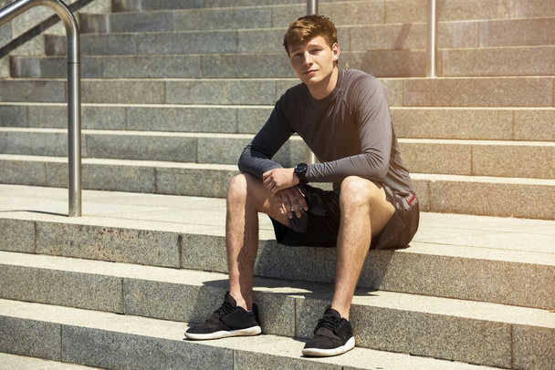 Young fit man resting after training outdoor, siting in full sun light in city. - Foto, immagini