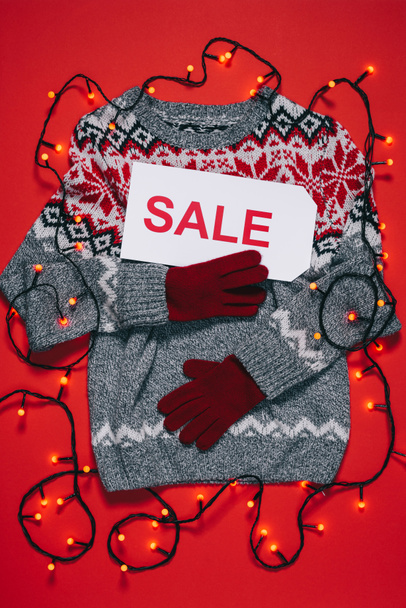 top view of winter sweater, mittens, price tag with sale lettering and christmas lights isolated on red - 写真・画像