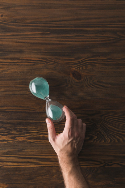 partial view of man holding hourglass in hand on wooden surface - Fotoğraf, Görsel