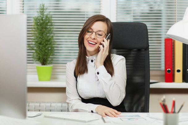 Mooie Glimlachende zakenvrouw in pak en bril zitten aan de balie, werkt op hedendaagse computer met documenten in lichte office, praten op mobiele telefoon, aangenaam gesprek voeren - Foto, afbeelding
