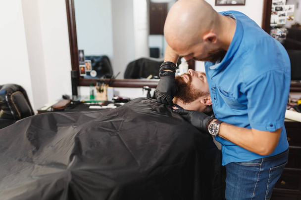 男性プロの美容師提供クライアントは、大きな、濃いひげストレートかみそりを剃るします。ジンジャー ハンサムなスタイリッシュな若い男短い髪、黒マントのトレンディなヘアカットを得るします。光白バーバー ショップ ルーム - 写真・画像