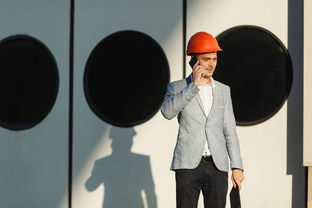 Young unshaven successful business man in gray suit and protective construction orange helmet holding tablet, talking on mobile phone, standing in sea port against doors with round windows background - Photo, Image