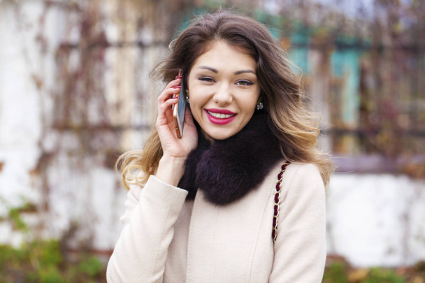 Portrait of happy young brunette woman in beige coat talking on  - Фото, изображение