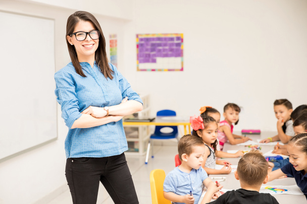 Nerdy Vorschullehrerin steht in einem Klassenzimmer voller Schüler und lächelt - Foto, Bild