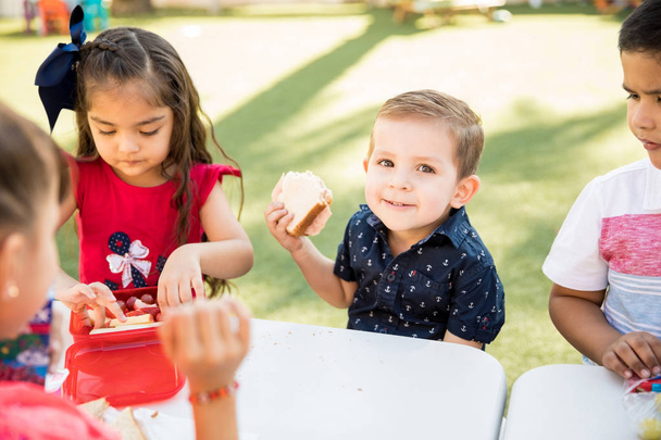Retrato de um menino comendo um sanduíche e sorrindo durante sua pausa para o almoço no jardim de infância
 - Foto, Imagem