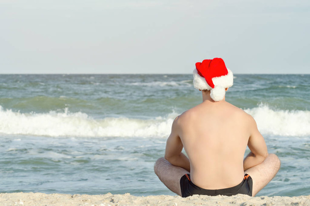 Ο άνθρωπος σε καπέλα Santa κάθεται στην παραλία. Πίσω όψη - Φωτογραφία, εικόνα