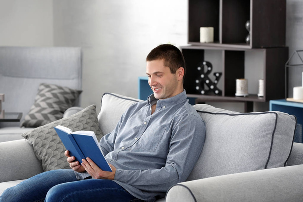 Young man reading book on sofa at home - 写真・画像