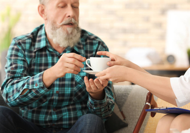 Young caregiver giving cup of tea to senior man at home - Photo, image