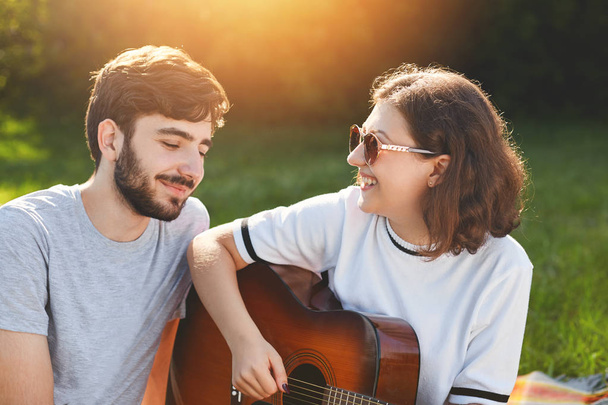 Outdoor shot of cheerful lovely couple spend free time on nature, play guitar and sign songs, look with love and cheerful expression at each other, enjoy beautiful nature and togetherness - Photo, image