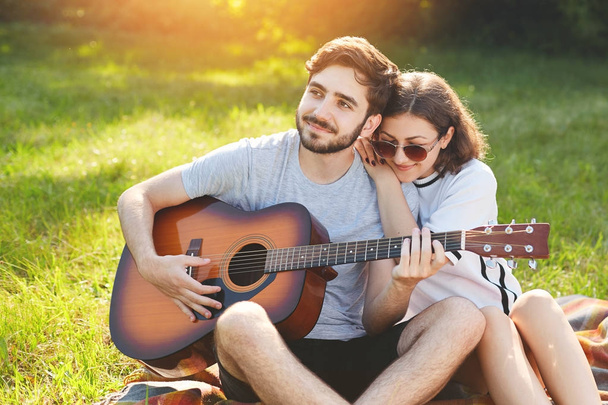 Dreamful romantic couple sit on green grass, enjoy togetherness. Handsome bearded man plays guitar and her girlfriend leans at his shoulder, listens to serenads. People and relationships concept - 写真・画像