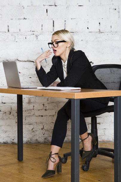 Joven mujer de negocios rubia de belleza sentada en una mesa de oficina con portátil, portátil y gafas de traje. Concepto empresarial
. - Foto, Imagen