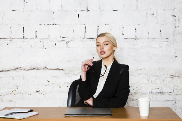 Молодая блондинка-бизнесвумен сидит за столом в офисе с ноутбуком, ноутбуком и очками в костюме. Концепция бизнеса
. - Фото, изображение