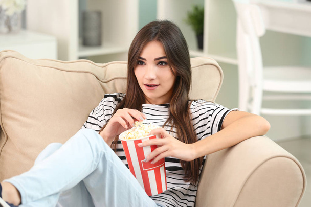 Young woman eating popcorn on sofa at home - Фото, изображение