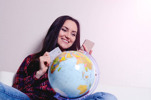 Happy woman with credit card and phone in hand, smiling sitting on sofa with globe. The joy of buying tickets for travel - Фото, изображение