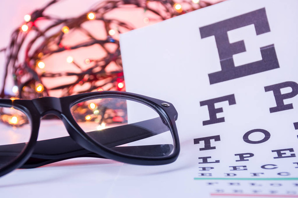 Weihnachten und Neujahr in der Augenoptometrie. Brille und ophthalmologischer Tisch für Sehschärfetest im Vordergrund mit verschwommenen Glühbirnen Weihnachtsgirlanden im Hintergrund - Foto, Bild