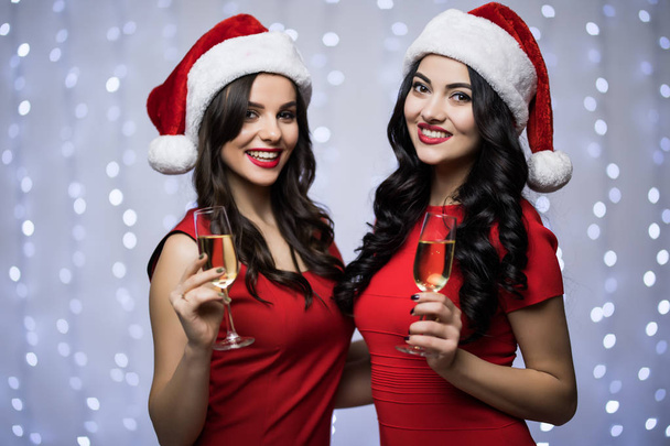 Porträt zweier Frauen in Weihnachtsmannmützen und rotem Kleid mit Champagner in den Händen auf hellem Bokeh-Hintergrund. Winterurlaub Weihnachten und Neujahr Konzept - Foto, Bild