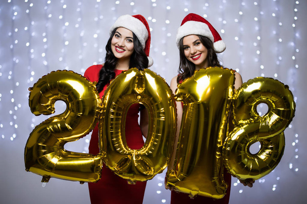 Retrato de dos mujeres en sombrero de santa y vestido rojo con números 2018 en las manos og Año Nuevo sobre fondo claro bokeh
. - Foto, imagen