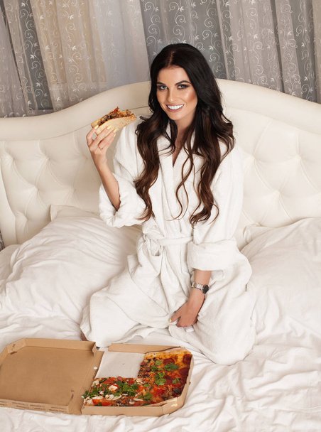 Pretty young sexy fashion sensual woman posing on white background.Sensual woman in white bathrobe. White bed isolated, crazy night party time,girl eating tasty pizza - Fotografie, Obrázek