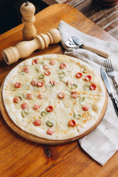 Hoge hoekmening van smakelijke pizza met salami en komkommers op houten trencher met linnen servet en zilverwerk - Foto, afbeelding