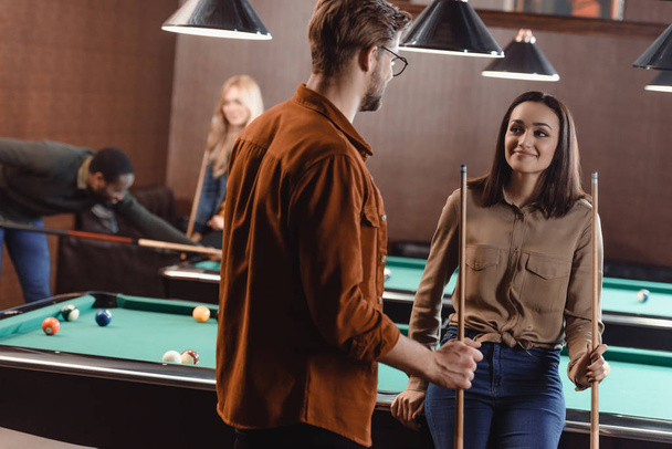 young man and woman standing beside pool table and looking at each other at bar with friends - Foto, immagini