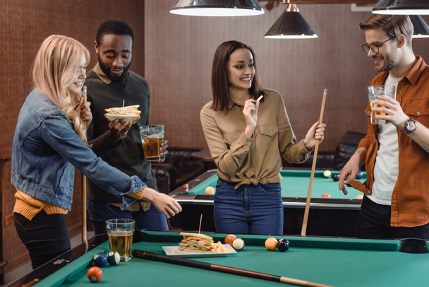 company of multiethnic friends eating and drinking beside pool table at bar  - Photo, Image