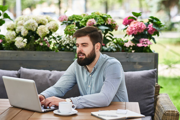 Портрет сучасного бородатого чоловіка, який використовує ноутбук за столом у зручному кафе на відкритому повітрі
 - Фото, зображення
