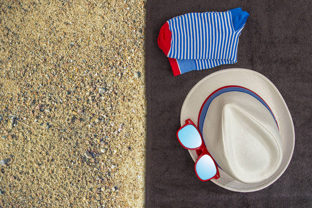 Мужской набор вещей для летнего отдыха (шляпа, солнцезащитные очки, носки) на темном полотенце и желтом песчаном фоне. Вид сверху
.  - Фото, изображение