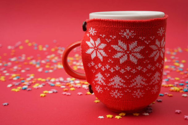 Cup of Hot Coffee or Tea  on the Table on red background. Cozy Festive Red Mug in knitted cover  with a Warm Drink. Christmas Morning Concept                 - Foto, afbeelding