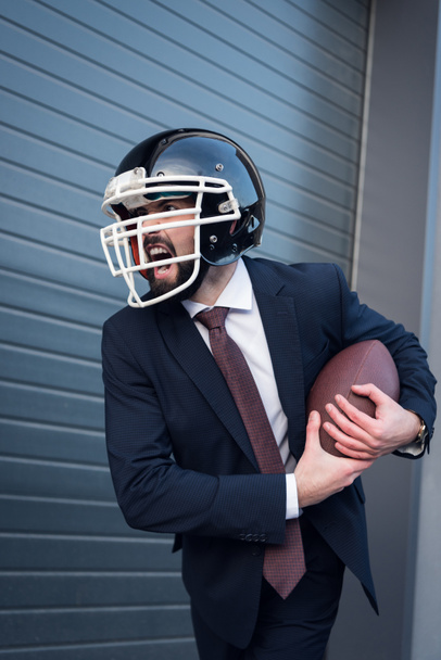 young angry businessman in suit and rugby helmet with ball in hands on street - Фото, зображення