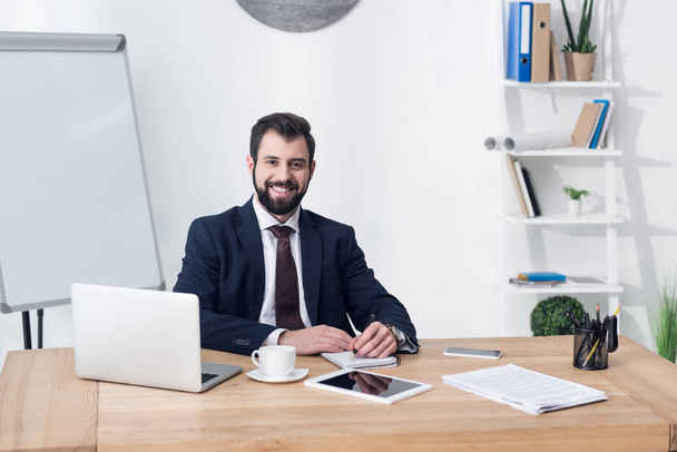 portrait of smiling businessman looking at camera while sitting at workplace in office - Photo, Image