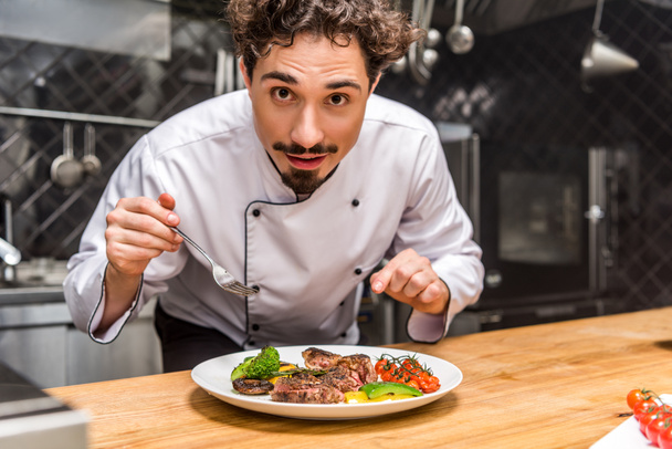 handsome chef standing with fork above cooked vegetables with meat - Φωτογραφία, εικόνα