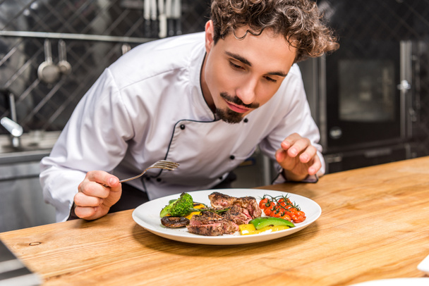 handsome chef standing with fork above fried vegetables with meat - Photo, Image