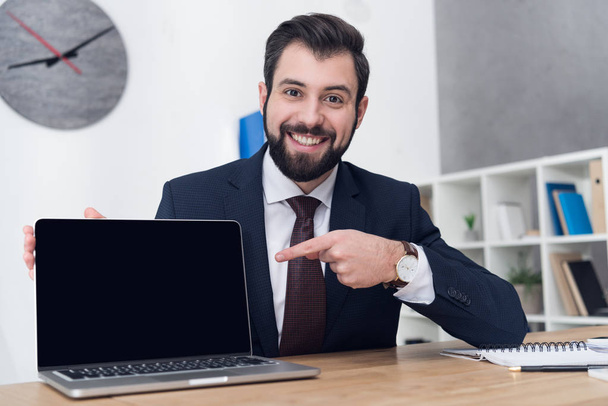 portrait of cheerful businessman pointing at laptop at workplace in office - Zdjęcie, obraz