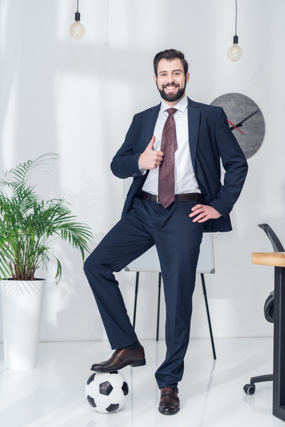 cheerful businessman standing with one leg on soccer ball and showing thumb up in office - Φωτογραφία, εικόνα