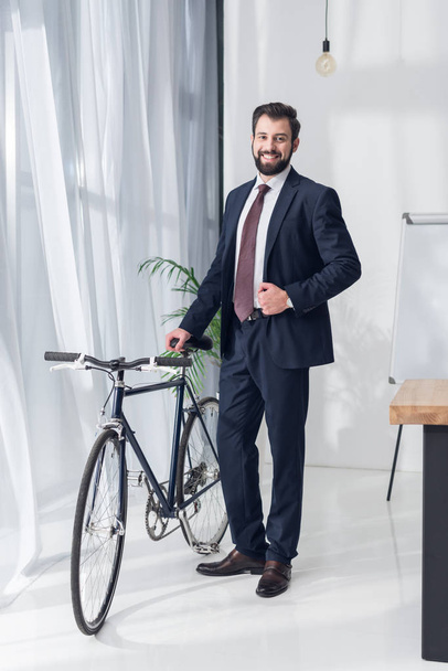 sonriente hombre de negocios en traje de pie cerca de la bicicleta en la oficina
 - Foto, imagen