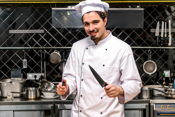 smiling chef standing with knifes in kitchen - Zdjęcie, obraz