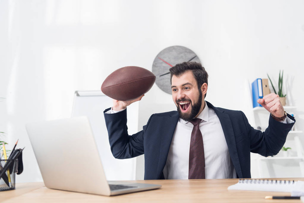 portrait of excited businessman with rugby ball at workplace with laptop in office - Foto, imagen